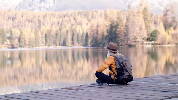 Rear view of senior woman hiker with hat sitting outdoors by lake in nature, resting. Slow motion.