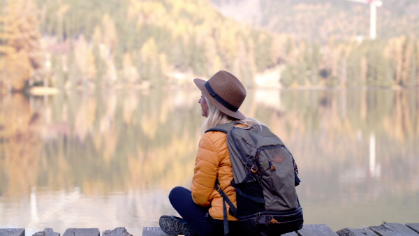 Rear view of senior woman hiker with hat sitting outdoors by lake in nature.