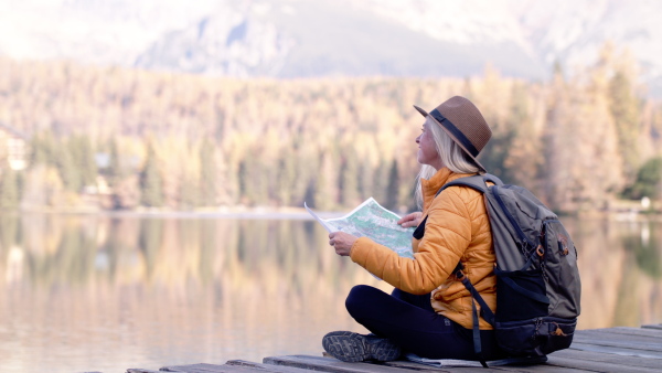 Active senior woman hiker with backpack sitting outdoors in nature, using map.