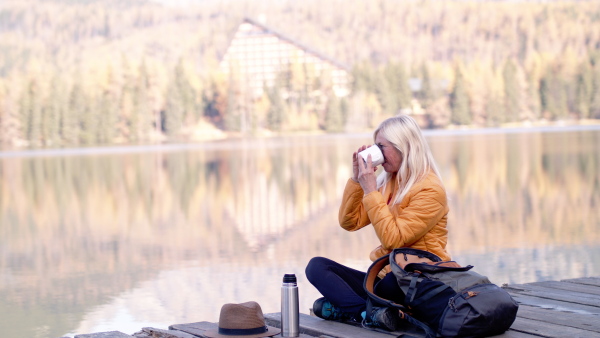 Side view of senior woman hiker sitting outdoors by lake in nature, drinking. Slow motion.