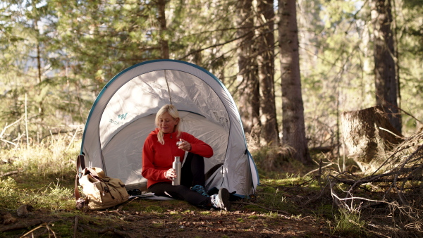 Senior woman hiker with tent shelter sitting outdoors in forest in nature, drinking.Slow motion.