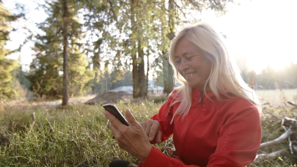 Active senior woman hiker sitting outdoors in nature, using smartphone. Slow motion.