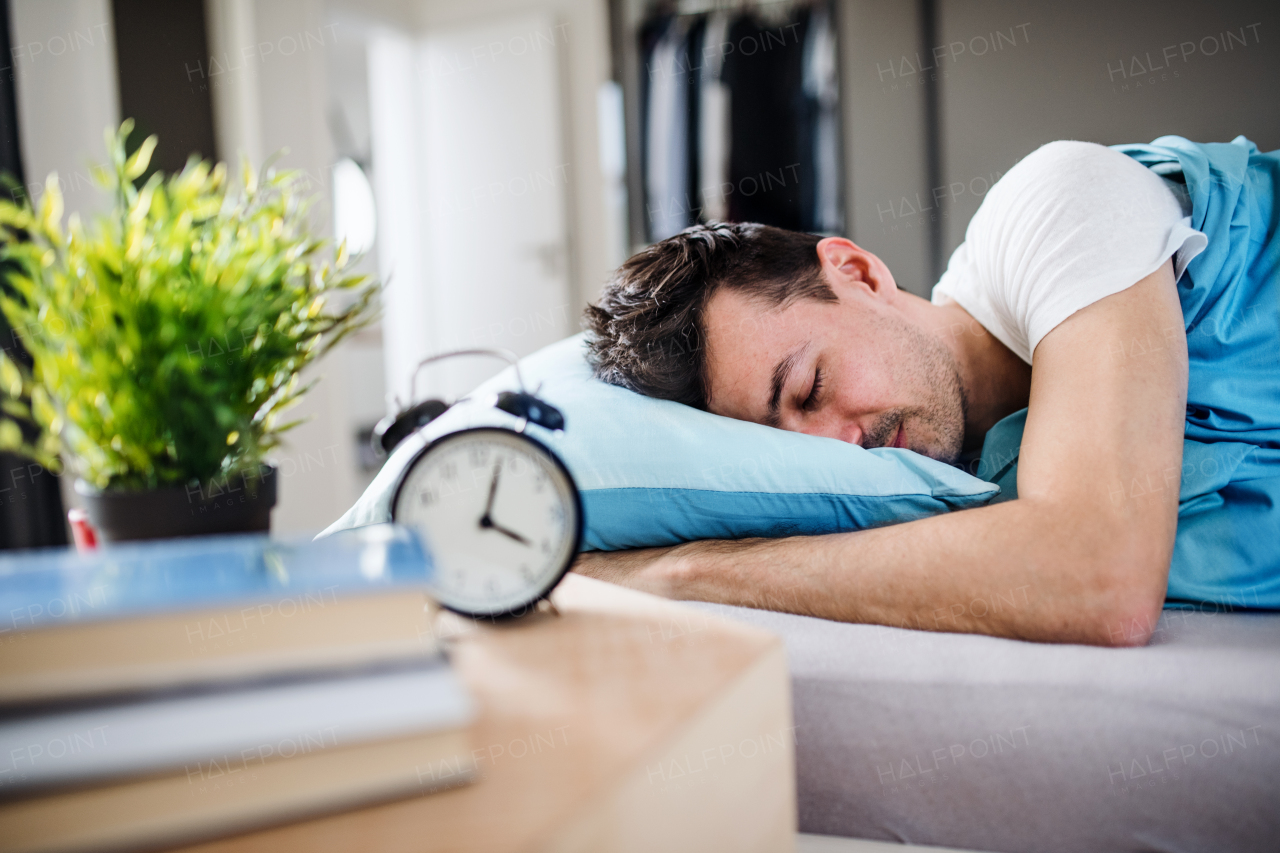 A young tired man with alarm clock in bed at home, sleeping.