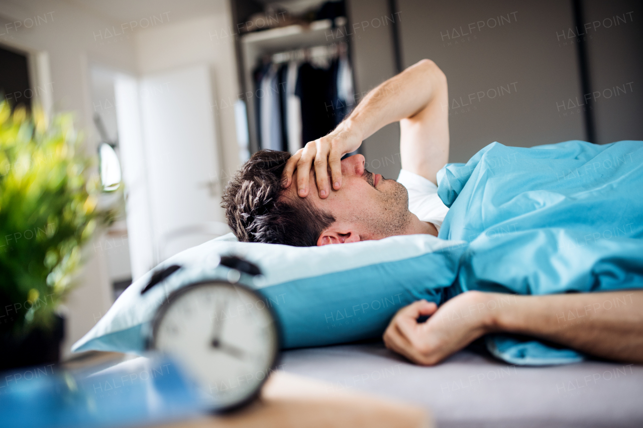 A young tired man with alarm clock in bed at home, waking up in morning.