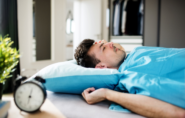 A young tired man with alarm clock in bed at home, sleeping.