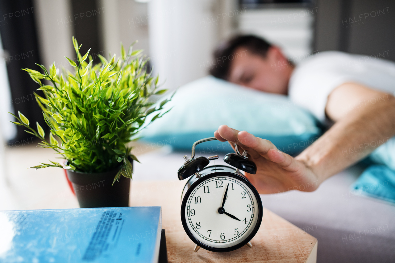 A young tired man hitting alarm clock in bed at home, waking up in morning.