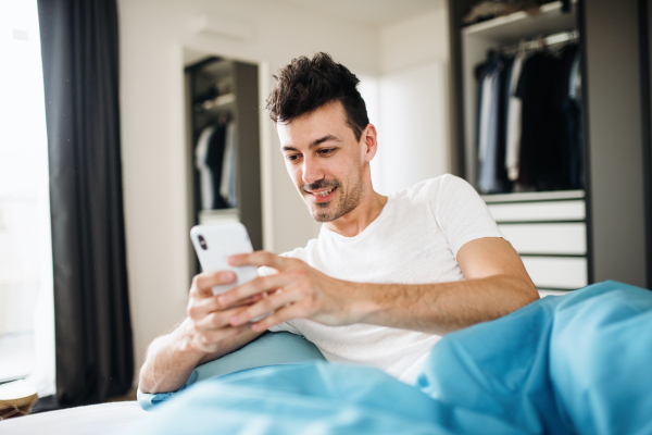 A young man with smartphone in bed at home, text messaging.