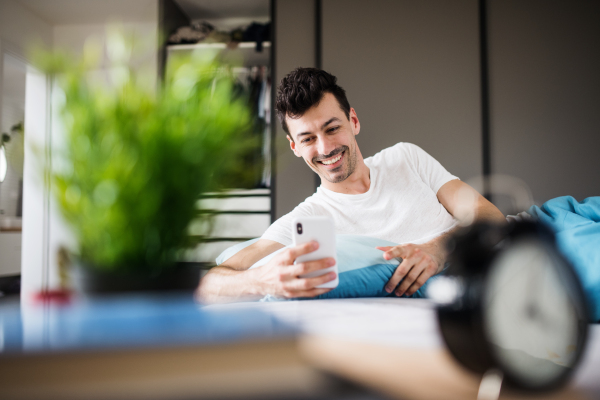 A young man with smartphone in bed at home, text messaging.