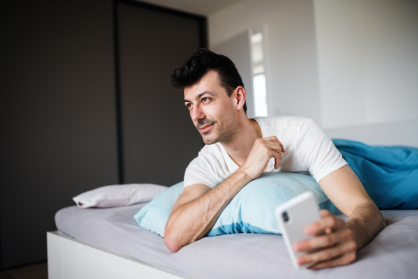 A young man with smartphone in bed at home, text messaging.