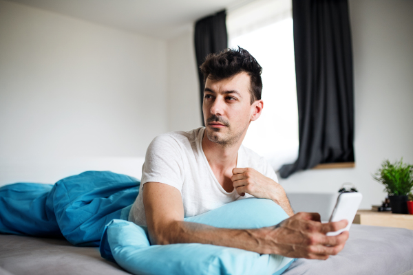 A young man with smartphone in bed at home, text messaging.