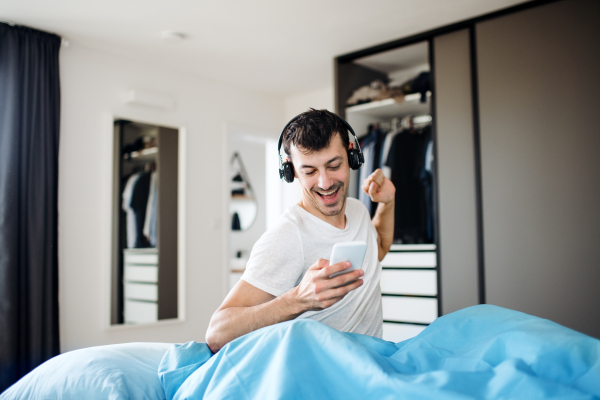 Young man with headphones and smartphone in bed indoors at home, listening to music.