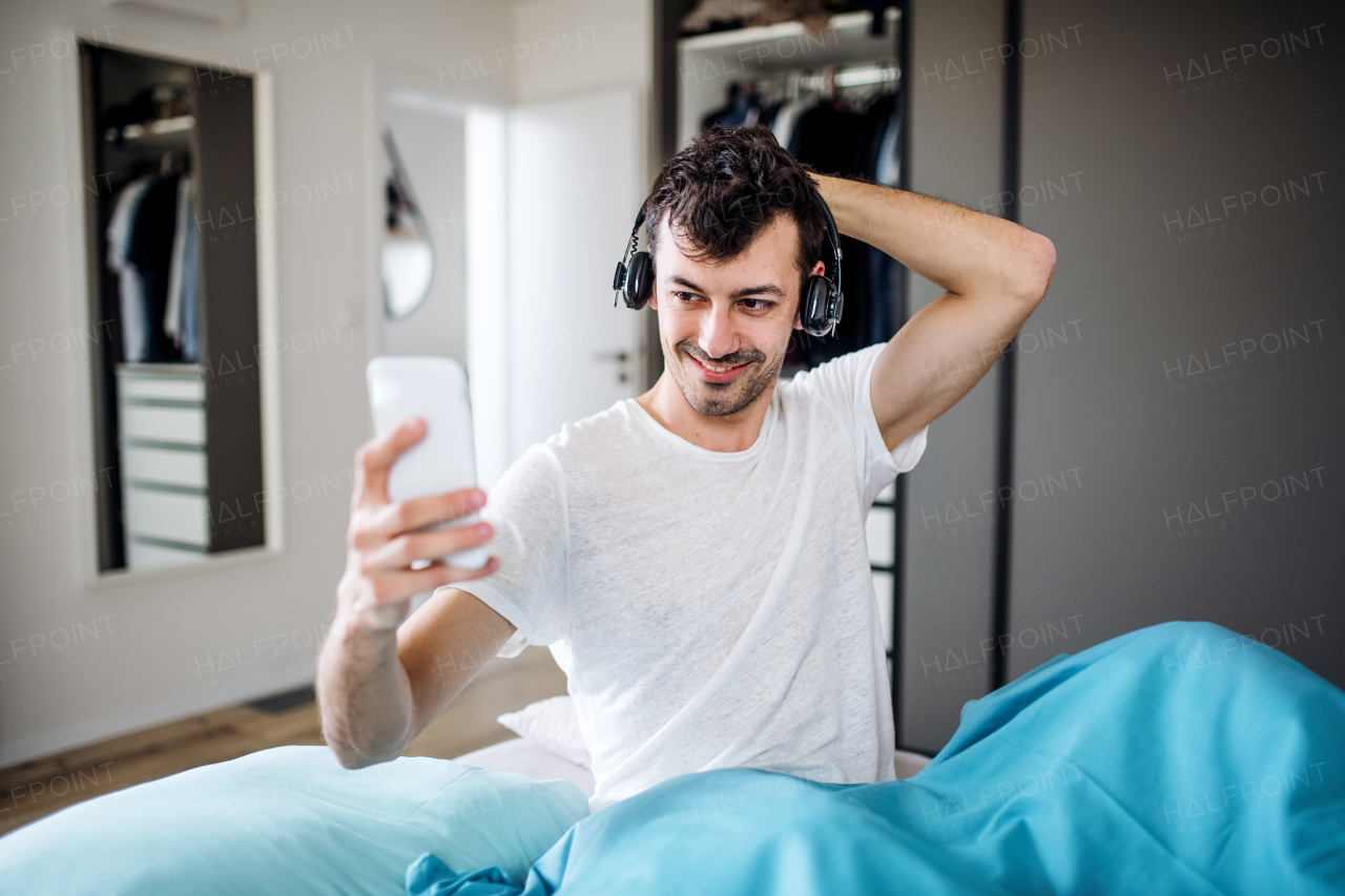 Young man with headphones and smartphone in bed indoors at home, listening to music.