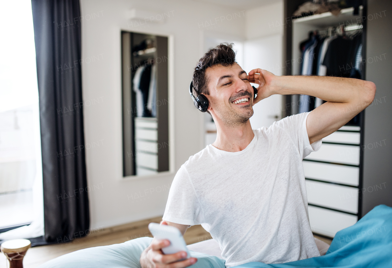 Young man with headphones and smartphone in bed indoors at home, listening to music.