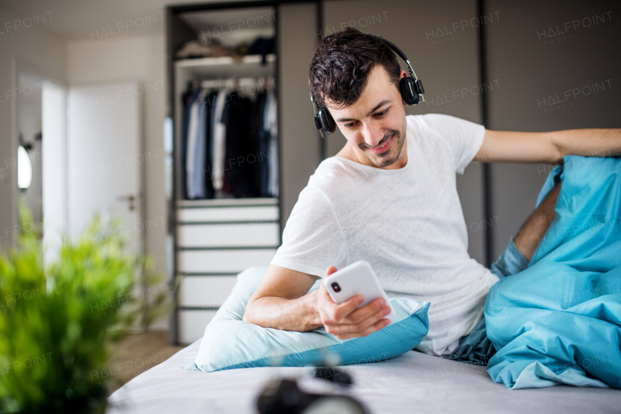 Young man with headphones and smartphone in bed indoors at home, listening to music.