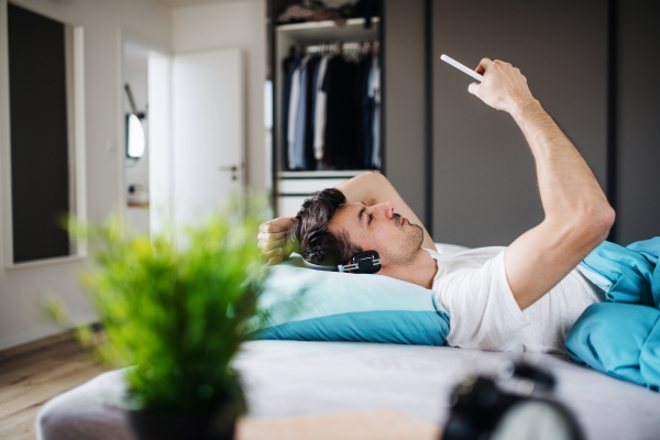 Young man with headphones and smartphone in bed indoors at home, listening to music.