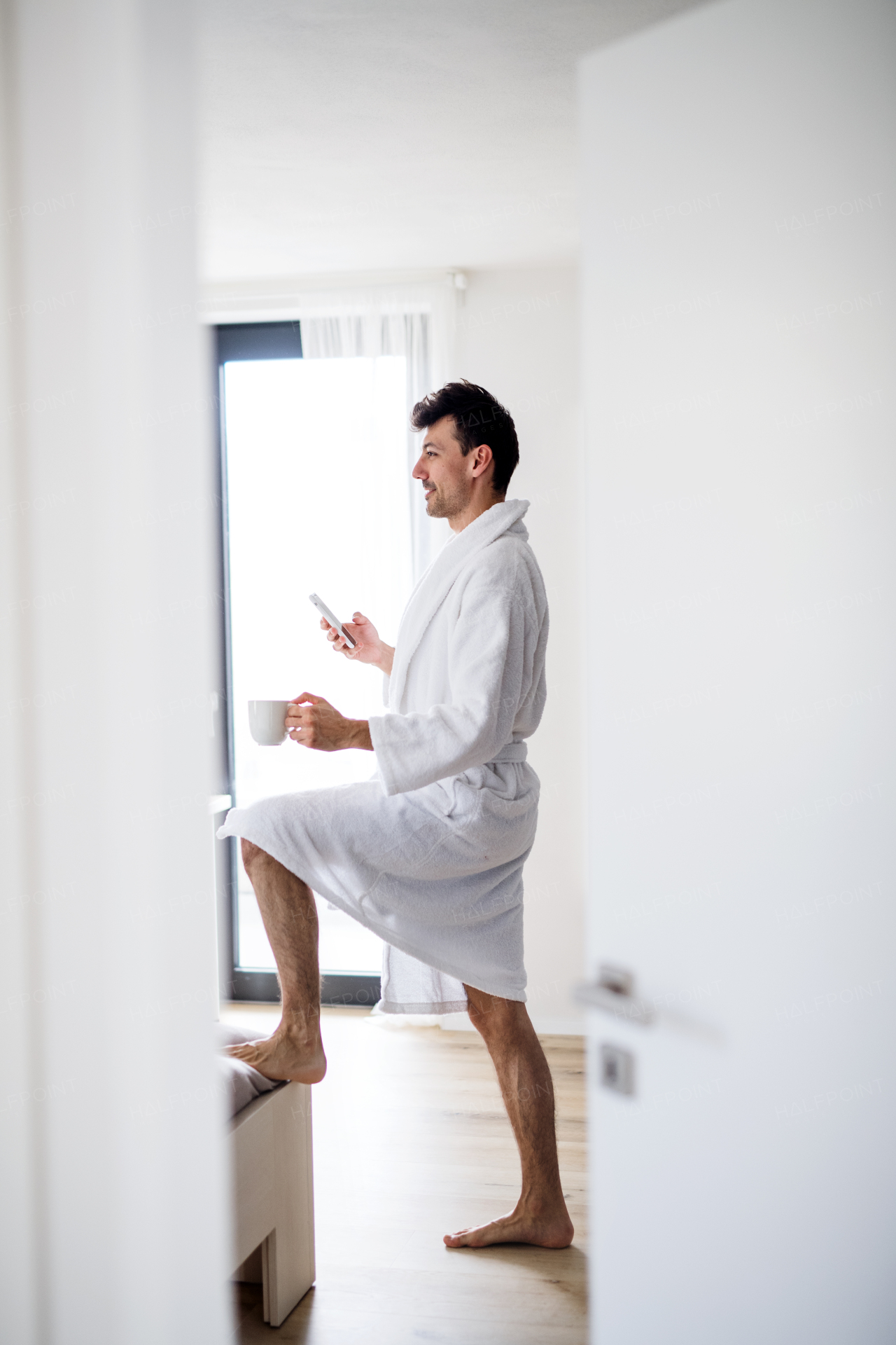 Young man with coffee standing in bedroom in the morning, using smartphone.