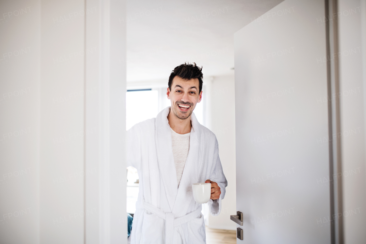 Front view of young man with coffee and dressing gown in bedroom in the morning.