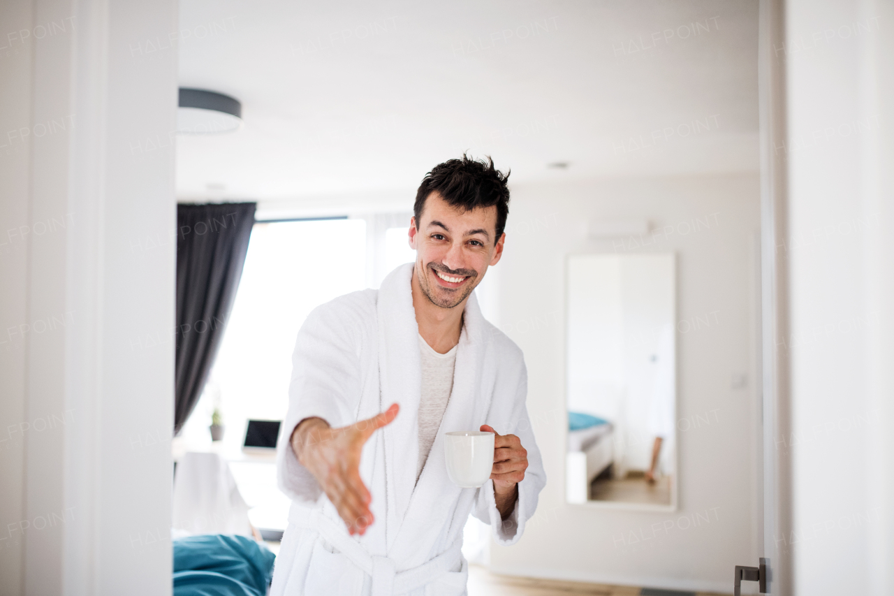 A young man with coffee offering hand for greeting in the bedroom, a morning routine.