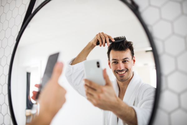 Young man combing hair in the bathroom in the morning, taking selfie. A daily routine.