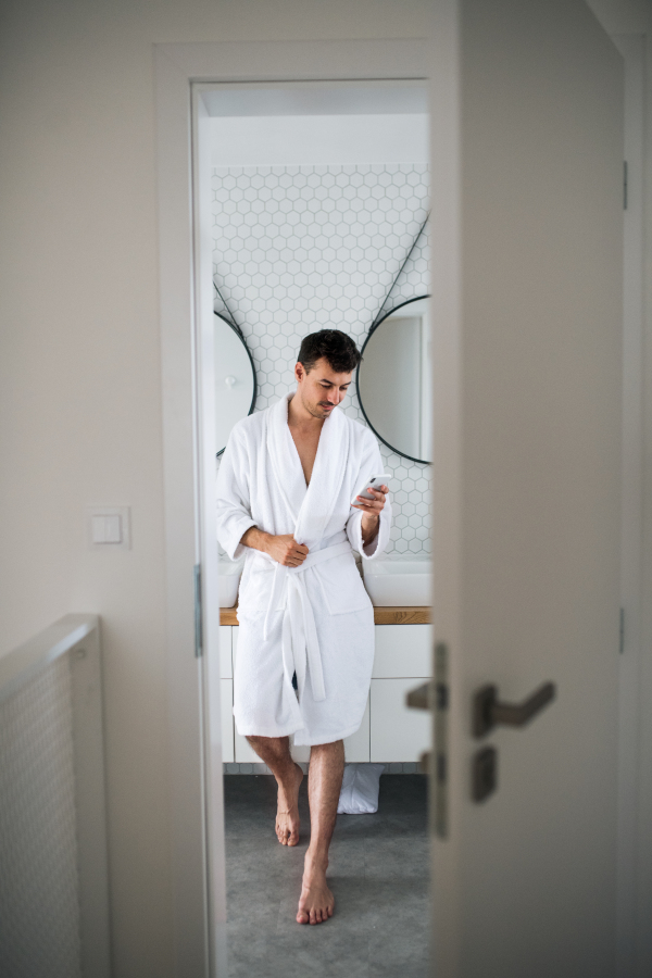 Young man standing in the bathroom in the morning, using smartphone.