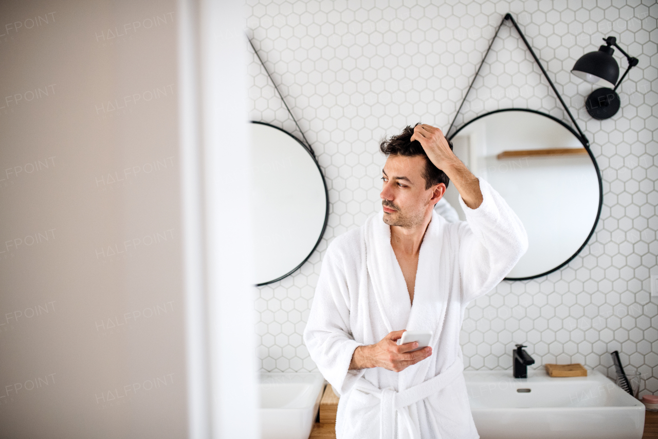 Young man standing in the bathroom in the morning, using smartphone.