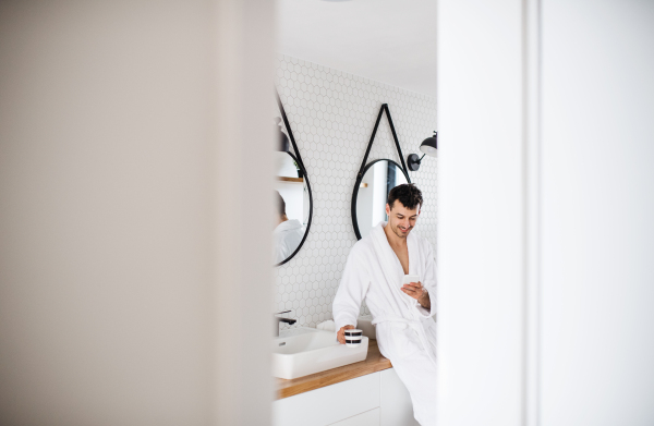 Young man standing in the bathroom in the morning, using smartphone.