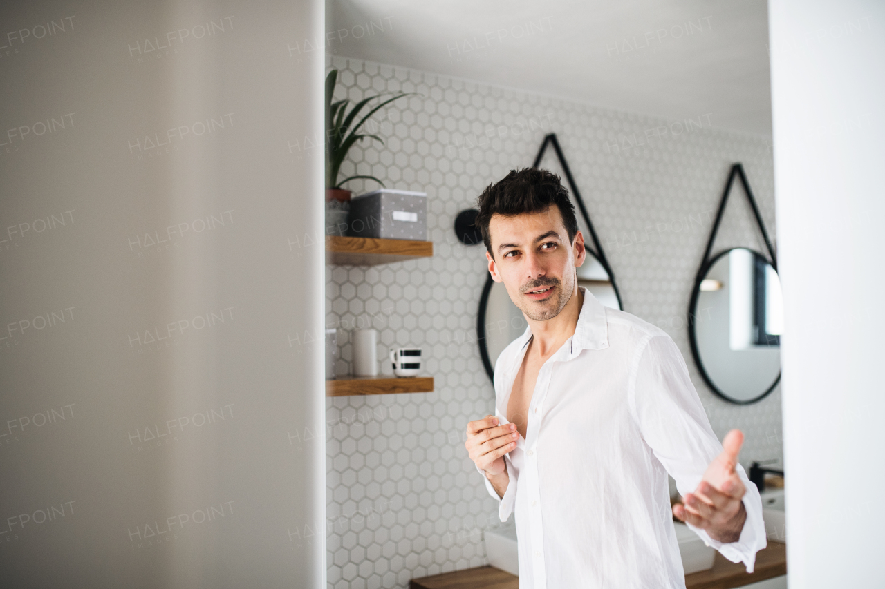 A young man putting on shirt in the bathroom in the morning.