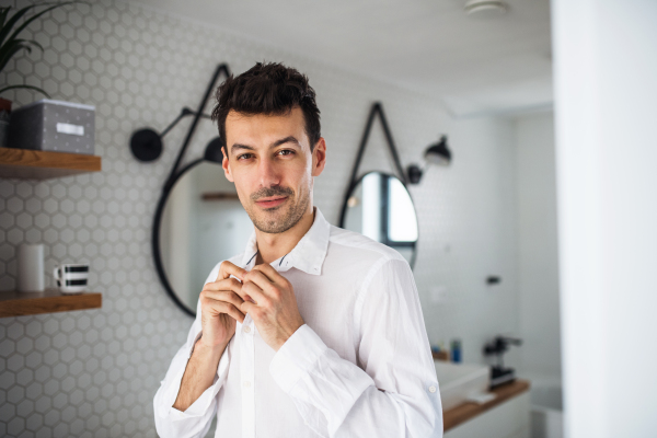 A young man putting on shirt in the bathroom in the morning.