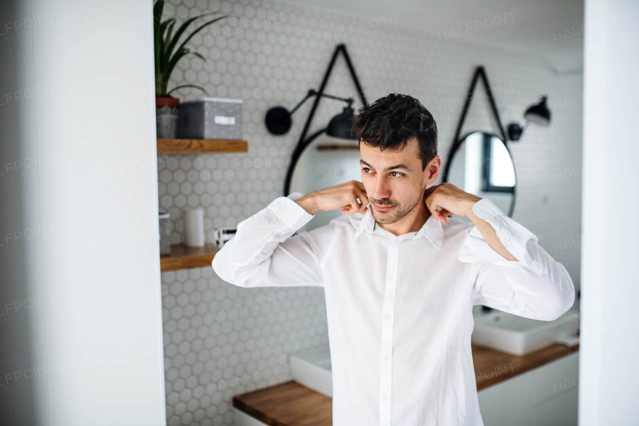 A young man putting on shirt in the bathroom in the morning.