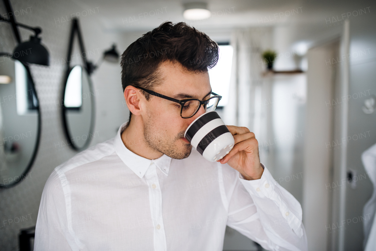 A young man with white shirt in the bathroom in the morning, drinking coffee.