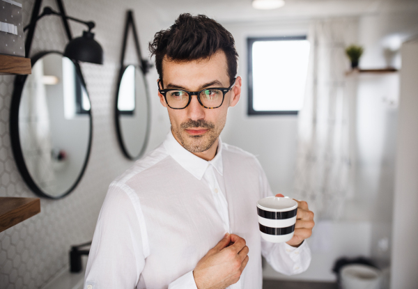 A young man with white shirt in the bathroom in the morning, holding a cup of coffee.