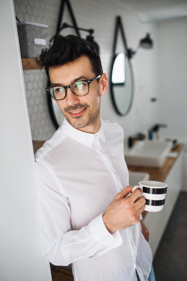 A young man with white shirt in the bathroom in the morning, holding a cup of coffee.