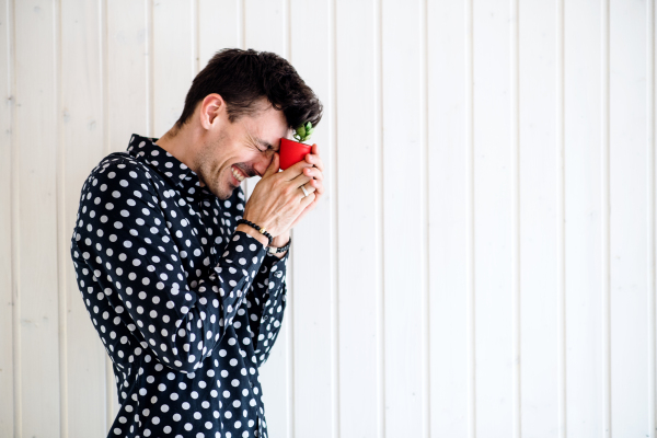 Frustrated young man standing against white wooden background wall, holding a small plant. Copy space.