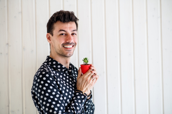 Young man standing against white wooden background wall, holding a small plant. Copy space.