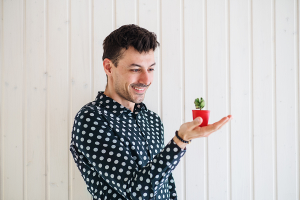 Young man standing against white wooden background wall, holding a small plant.