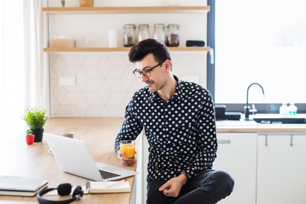 Young man with laptop sitting in kitchen, holding juice. A home office concept.