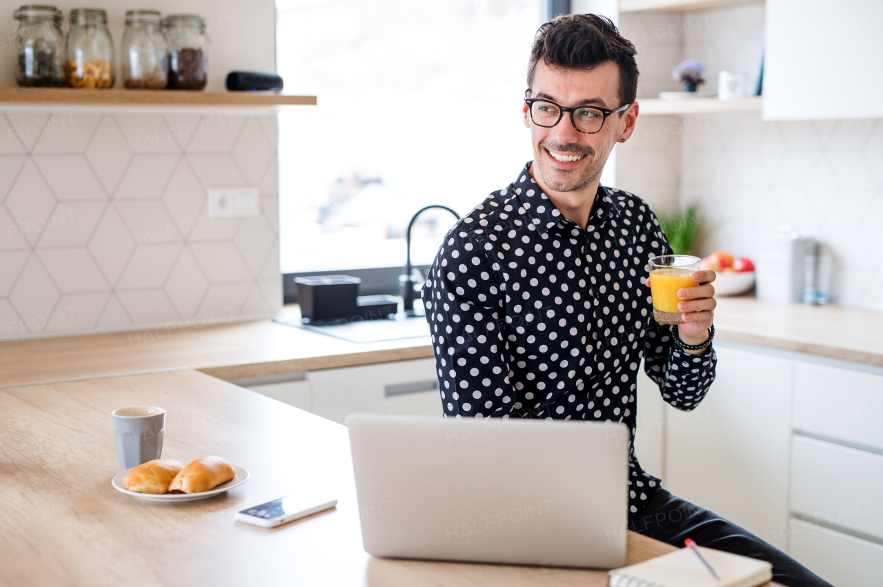 Young man with laptop sitting in kitchen, holding juice. A home office concept.