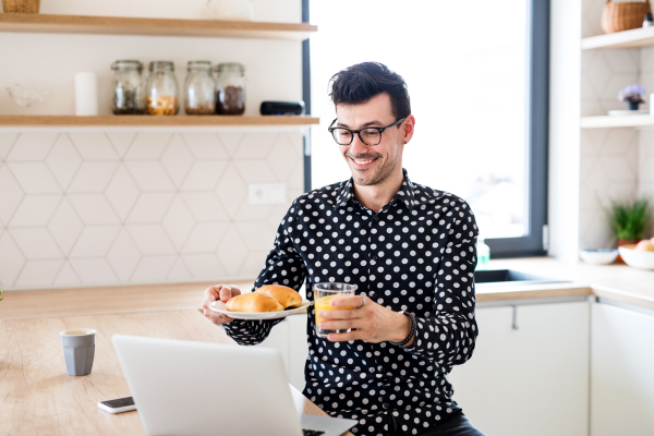 Cheerful young man with breakfaset and laptop indoors at home having skype call.