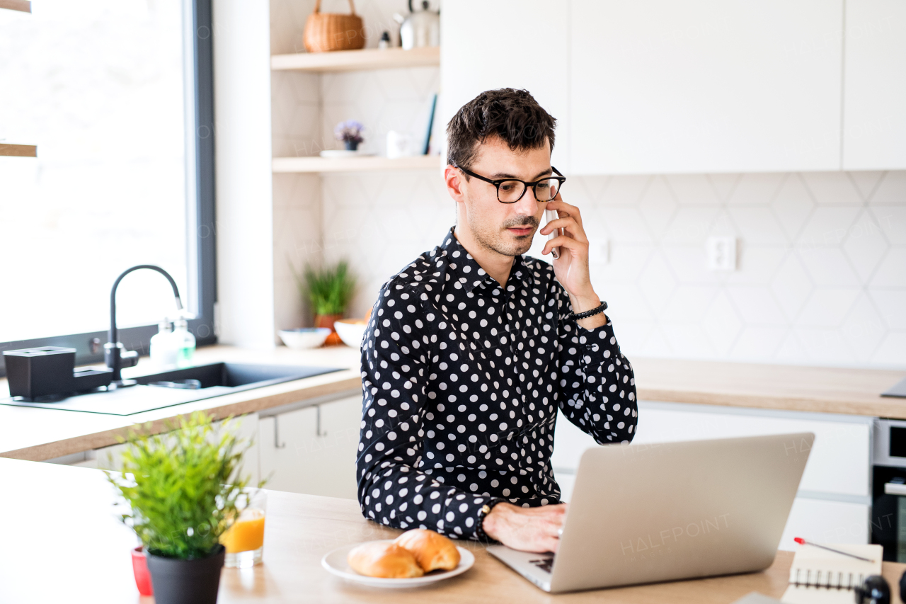 Young man with laptop and smartphone sitting in kitchen, working. A home office concept.