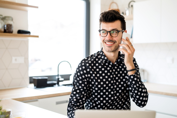 Young man with laptop and smartphone sitting in kitchen, making a phone call. A home office concept.