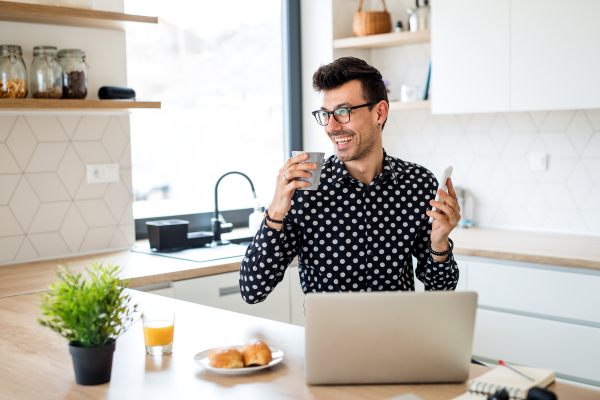 Young man with laptop and smartphone sitting in kitchen, working. A home office concept.