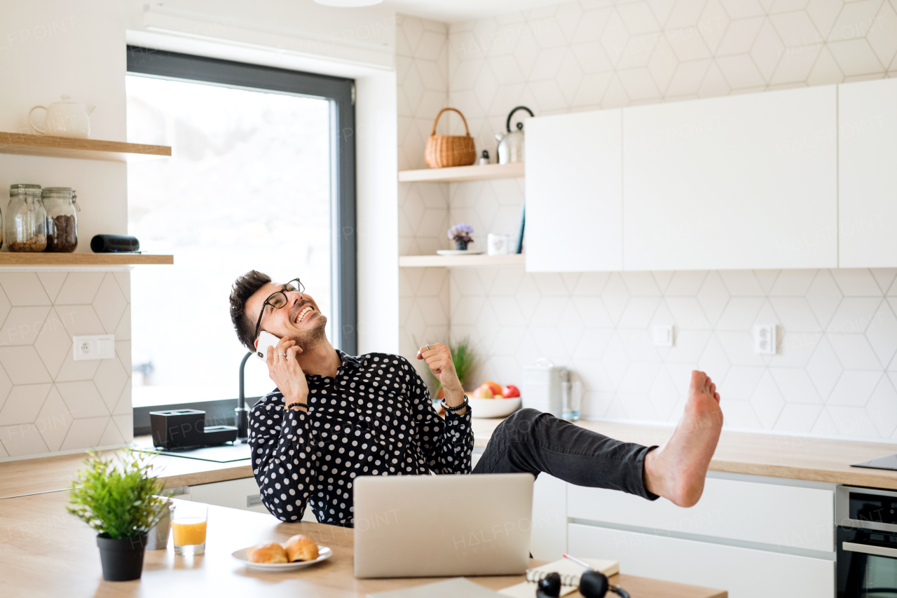 Cheerful young man with laptop and smartphone sitting in kitchen, expressing excitement when making a phone call. A home office concept.