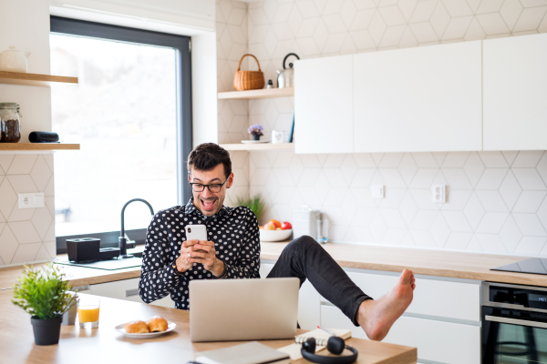 A young man with smartphone and laptop, eating breakfast indoors at home.