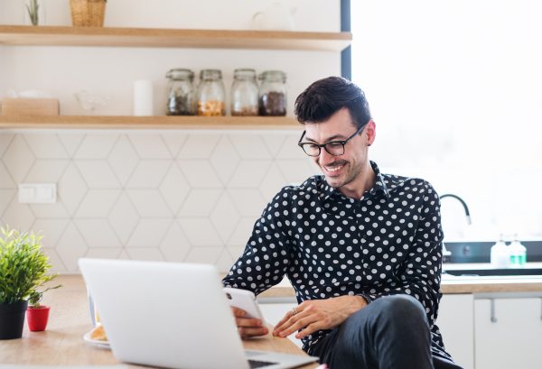 Young man with laptop and smartphone sitting in kitchen, texting. A home office concept.