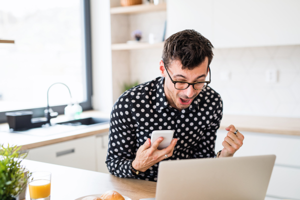 Young man with laptop and smartphone sitting in kitchen, expressing excitement. A home office concept.