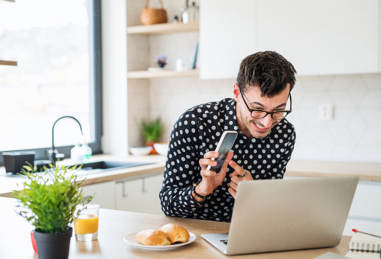 Young man with laptop and smartphone sitting in kitchen, working. A home office concept.