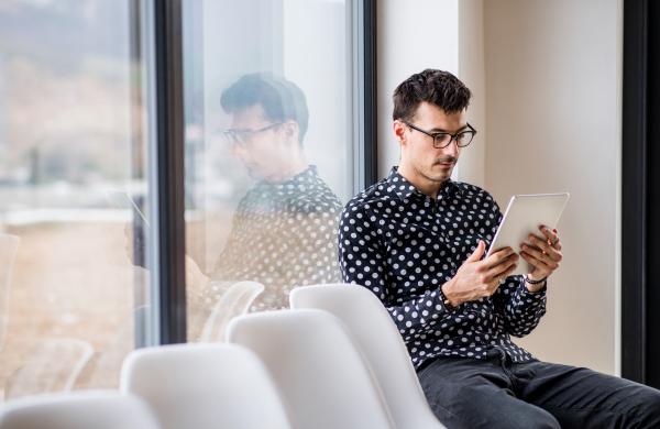 A young man with tablet sitting by the window in waiting room. Copy space.