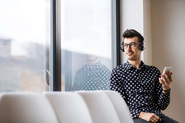 A young man with headphones and smartphone sitting by the window, listening to music.