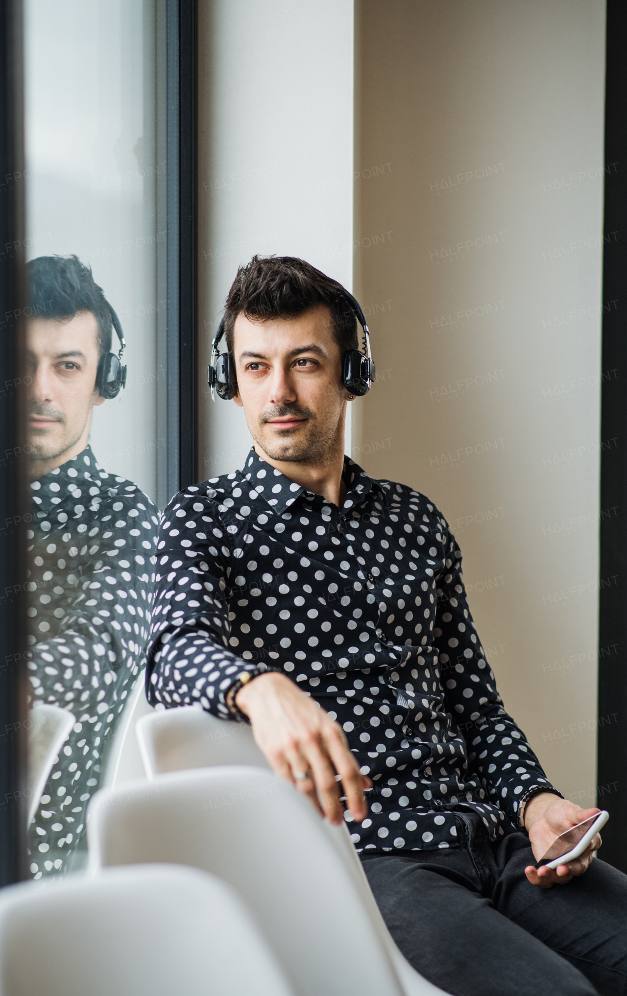 A young man with headphones and smartphone sitting by the window, listening to music.