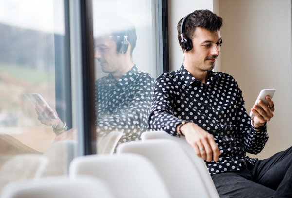 A young man with headphones and smartphone sitting by the window, listening to music.